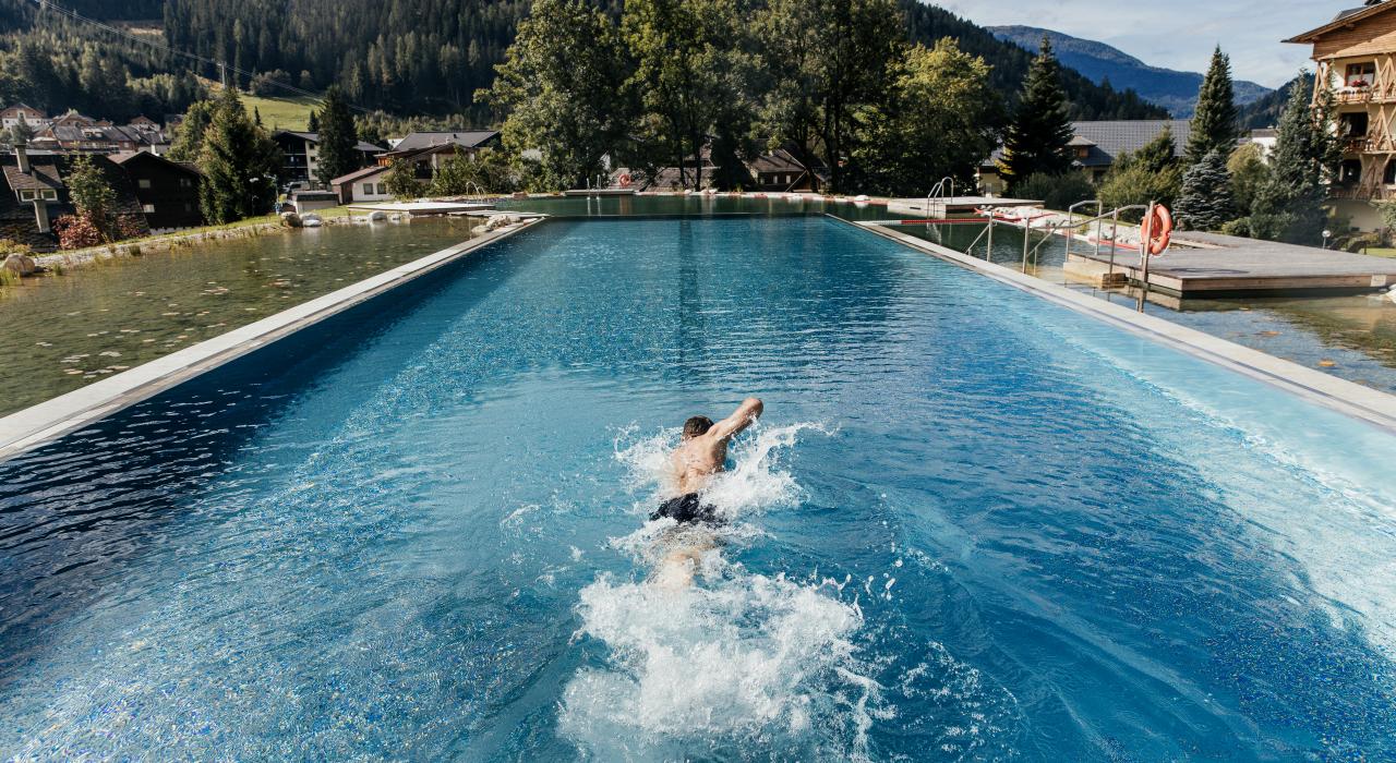 Ein Schwimmer zieht seine Bahnen in einem beeindruckenden Außenpool, umgeben von einer malerischen Berglandschaft. Die üppigen Bäume und sanften Hügel bieten eine ruhige Kulisse, während das klare Wasser das Licht der Sonne reflektiert.