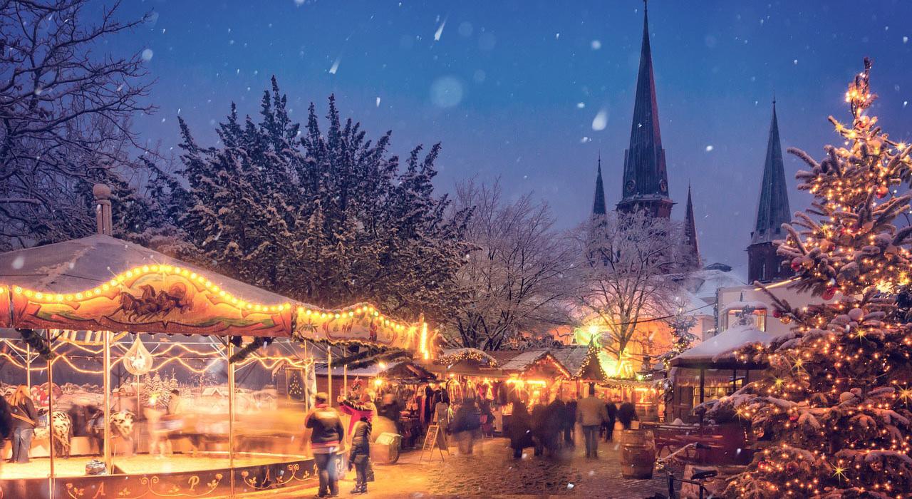 Ein Weihnachtsmarkt mit Karussell und Leuten, die vorbei laufen, im Hintergrund eine Kirche