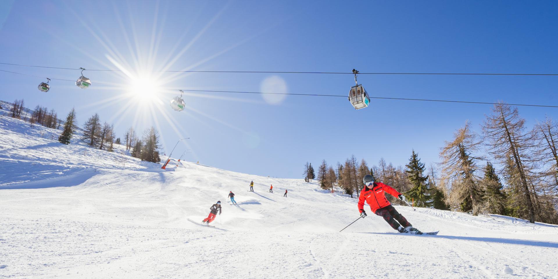 Mehrere Skifahrer fahren eine sonnige Skipiste hinunter, während eine Seilbahn über ihnen verläuft