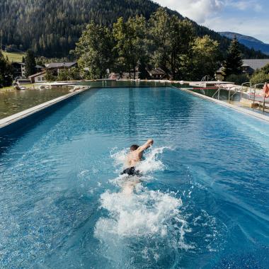 Ein Schwimmer zieht seine Bahnen in einem beeindruckenden Außenpool, umgeben von einer malerischen Berglandschaft. Die üppigen Bäume und sanften Hügel bieten eine ruhige Kulisse, während das klare Wasser das Licht der Sonne reflektiert.