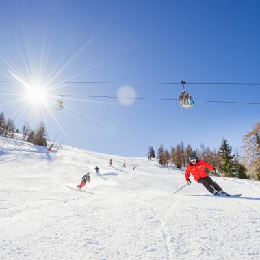 Mehrere Skifahrer fahren eine sonnige Skipiste hinunter, während eine Seilbahn über ihnen verläuft