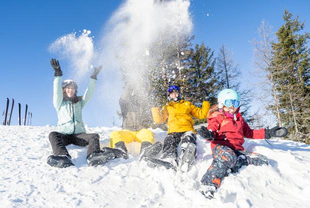 Eine Gruppe Menschen in Winterkleidung sitzt im Schnee und wirft lachend Schneebälle in die Luft