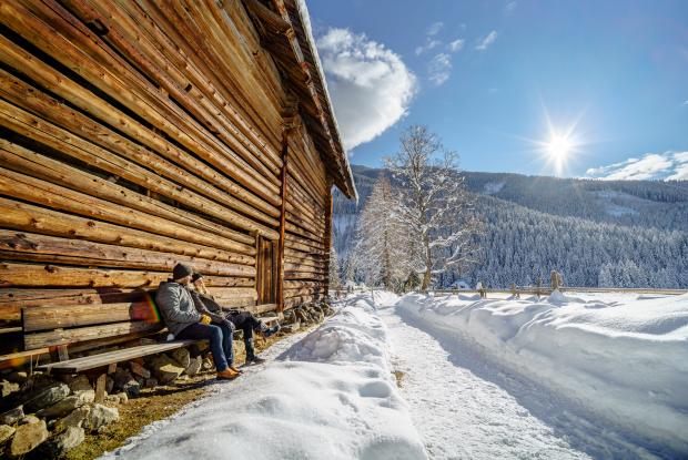 Zwei Menschen sitzen neben einer Holzhütte und genießen die verschneite Natur während ihres Winterurlaubs in Kärnten