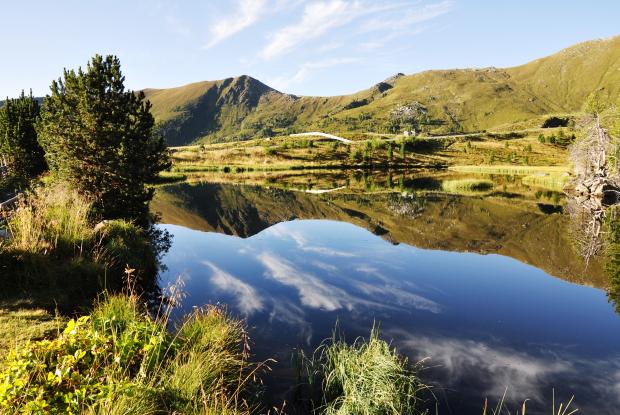 Ein ruhiger Bergsee, umgeben von grünen Hügeln und Bergen, spiegelt den klaren Himmel wider