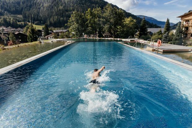 Ein Schwimmer zieht seine Bahnen in einem beeindruckenden Außenpool, umgeben von einer malerischen Berglandschaft. Die üppigen Bäume und sanften Hügel bieten eine ruhige Kulisse, während das klare Wasser das Licht der Sonne reflektiert.
